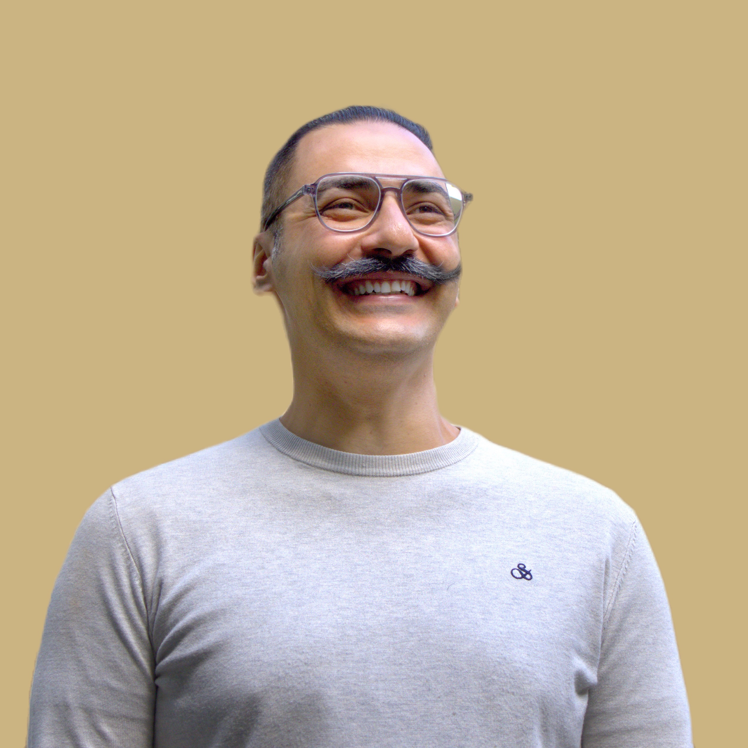 Aram, a man with a mustache and glasses sporting a smile standing in front of a building